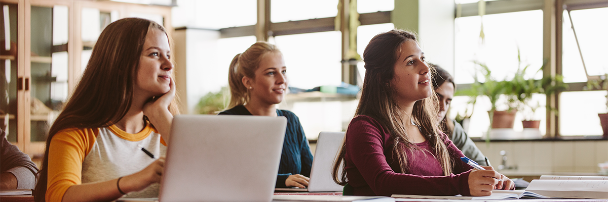 four students sitting in class taking notes on paper and laptop